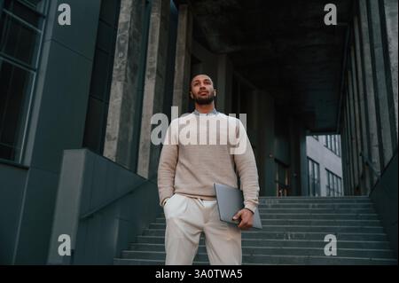 Front view. Standing with laptop in hands. Handsome black man is outdoors near the business building. Stock Photo