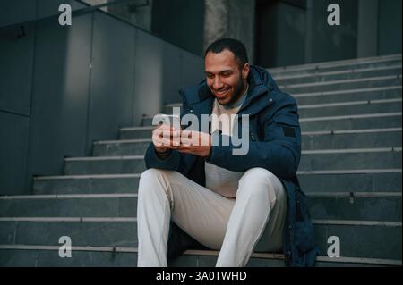 Sitting on the stairs with smartphone in hands. Handsome black man is outdoors near the business building. Stock Photo