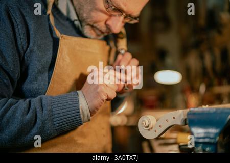 Cremona, Italy - February 6th 2025 Maestro Luca Tenore Italian luthier wearing an apron is using small tools to carefully carve the details of a violi Stock Photo