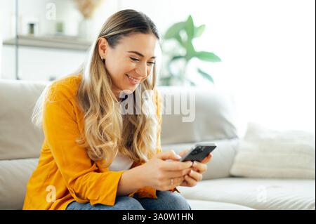 Pretty caucasian woman is sitting on a sofa, smiling as she looks at her phone, she seems happy, enjoying her personal leisure time while using her smartphone messaging online in a cozy living room Stock Photo