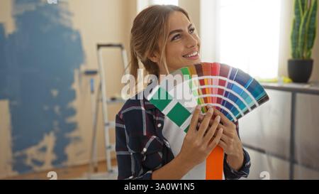Woman smiling in living room holding paint swatches for home renovation with blonde hair wearing plaid shirt and ladder visible in cozy apartment inte Stock Photo