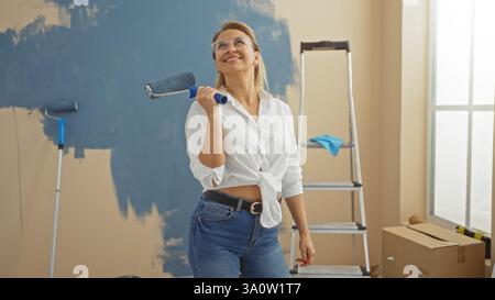 Woman painting wall indoors with roller wearing casual clothes in new home showcasing blonde hair and joyful expression in modern living room setting. Stock Photo
