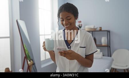 Woman smiling indoors holding a cup and smartphone in a wellness spa room with a mirror and shelves in the background Stock Photo