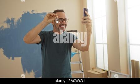 Man holding keys and smiling while taking a selfie in a newly painted living room, suggesting the joy of moving into a new home with bright sunlight f Stock Photo