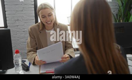 Two businesswomen sitting in an office discussing work documents, showcasing collaboration and teamwork in a professional workplace environment with c Stock Photo