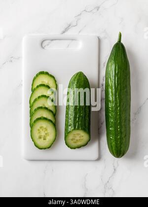 Raw green cucumbers on a white cutting board. Whole long cucumber, cut in half and juicy slices over marble countertop. Organic vegetables, harvest. Stock Photo