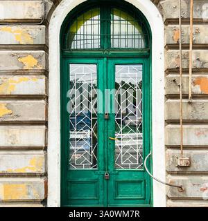 A weathered green door with iglass and metalwork, framed by the peeling facade of a historic building in Łódź Stock Photo