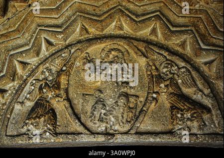 Christ in Majesty or Maiestas Domini on Norman Romanesque tympanum, studded with prehistoric sea shells embedded in sedimentary rock, Church of St Andrew, Eastleach, Gloucestershire, England. Sculpted figure of Jesus with cruciform halo sits on rectangular throne, right hand raised in gesture of blessing and left holding book.  Two winged angels support central mandorla below rounded arch and bands of typically Romanesque zigzag chevron moulding. Stock Photo