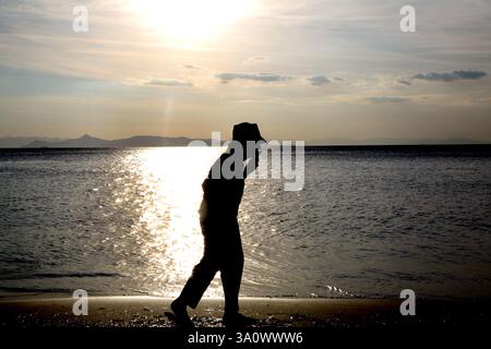 Silhouette of Woman Walking along shoreline Using Mobile Phone Vouliagmeni Beach Athens Attica Greece Stock Photo