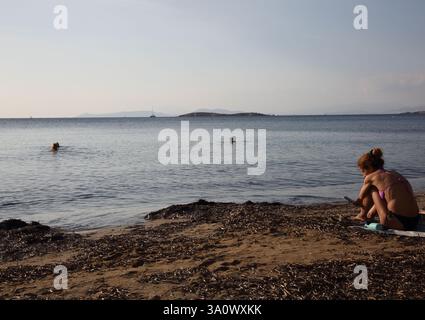 Woman Looking at Mobile Phone sitting on Vouliagmeni Beach Greece Stock Photo