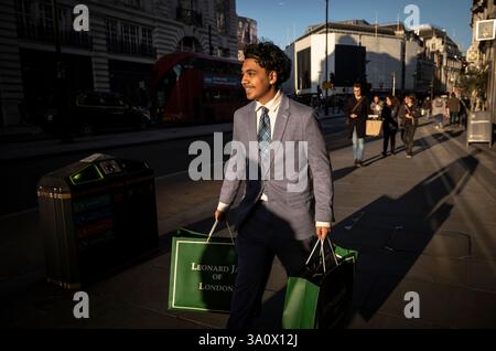 A young man carries clothes bags from Leonard Jay along Piccadilly, gentleman's outfitters based in the City of London, England, United Kingdom Stock Photo