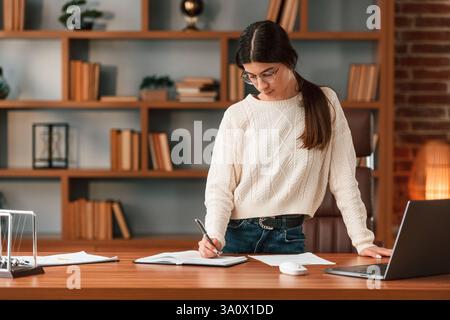 Young woman in glasses is working indoors. Standing by table with laptop. Stock Photo