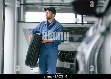 With tire in the hands. Man in blue uniform is working in the car service. Stock Photo