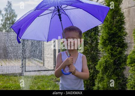Happy boy with a playful smile holding a purple umbrella in the backyard during summer, standing under a garden sprinkler Stock Photo