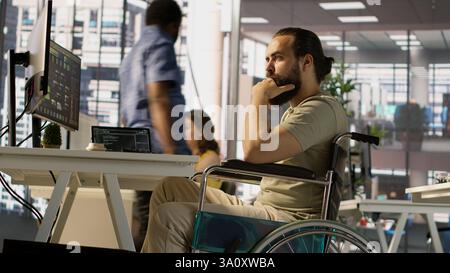 IT worker with disability sitting in wheelchair in office working at computer desk, focused on testing programs. Software engineer with paraplegia in inclusive workplace checking code on PC, camera A Stock Photo