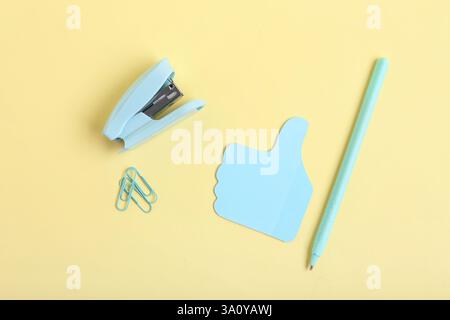 Blue stapler with clips, pen and sticky note in shape of thumb-up on yellow background Stock Photo