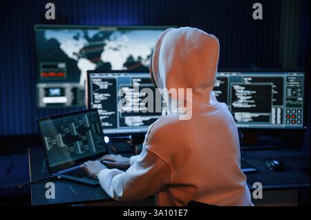 In white hoodie. Young professional female hacker is indoors by computer with lot of information on displays. Stock Photo
