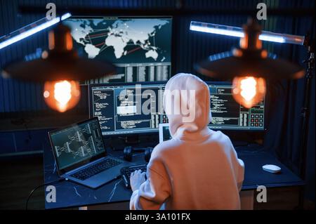 Sitting in white hoodie. Young professional female hacker is indoors by computer with lot of information on displays. Stock Photo