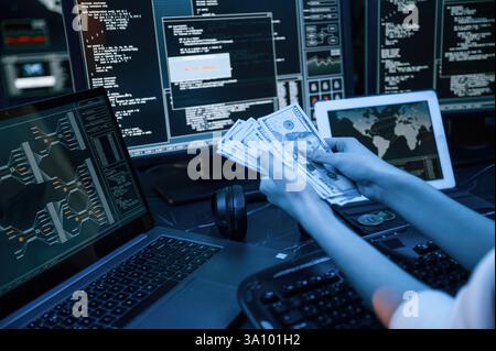 With currency in hands. Young professional female hacker is indoors by computer with lot of information on displays. Stock Photo