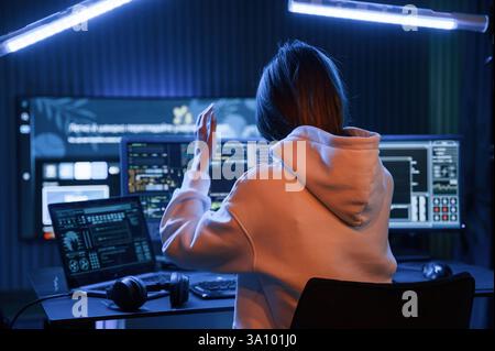 Looking at screens. Young professional female hacker is indoors by computer with lot of information on displays. Stock Photo