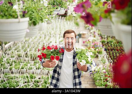 Holding flowers, smiling, positive person. Man is working in garden center. Successful employee is in a bright greenhouse. Stock Photo