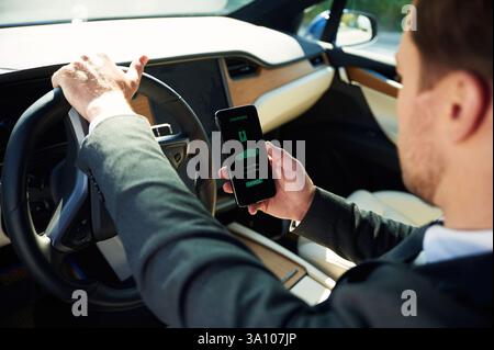Holding smartphone with charging progress app. Businessman in suit is driving his electric car. Stock Photo