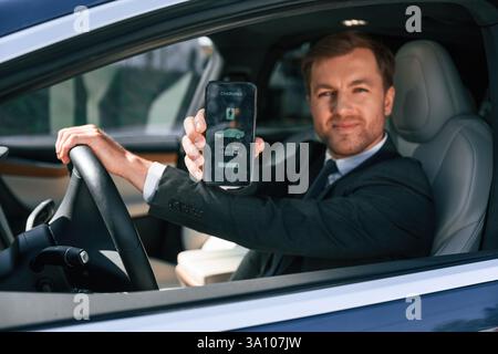 Holding smartphone with charging progress app. Businessman in suit is driving his electric car. Stock Photo