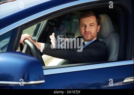 Holding smartphone with charging progress app. Businessman in suit is driving his electric car. Stock Photo