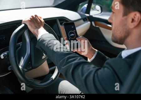Holding smartphone with charging progress app. Businessman in suit is driving his electric car. Stock Photo