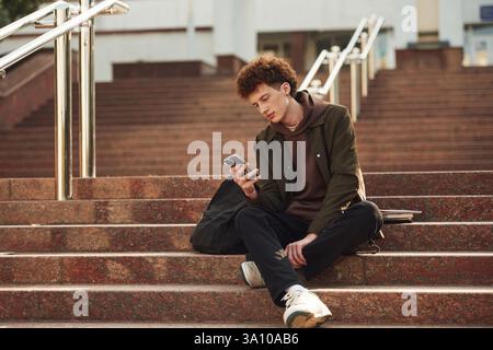 Smartphone is in hands. Young student with curly hair is outdoors near university. Stock Photo