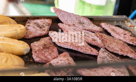 Thin slices of salamini cooking on a hot grill with visible charring while fresh bread rolls are placed nearby in preparation for making panini at an Stock Photo