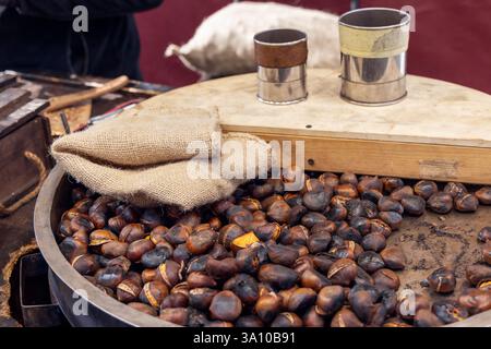 Metal roasting pan filled with freshly roasted chestnuts burlap sacks rest on top while metal measuring cups sit on a wooden board in a traditional st Stock Photo