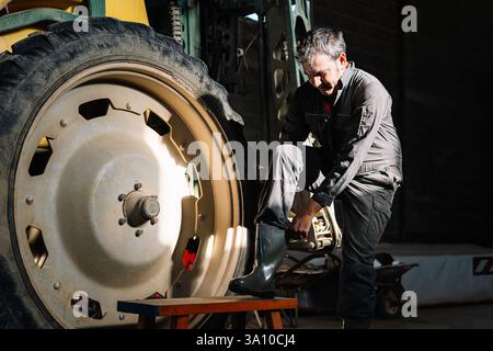 Caucasian male farmer putting on rubber boots next to a tractor wheel, getting ready for work in a barn Stock Photo