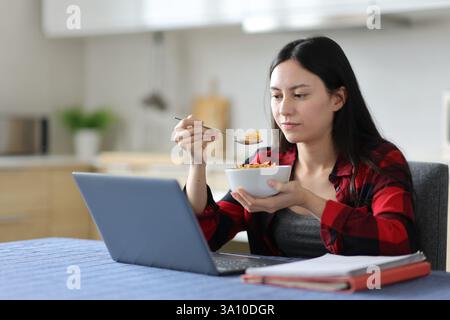Asian student eating cereals using laptop sitting in the kitchan in a house Stock Photo