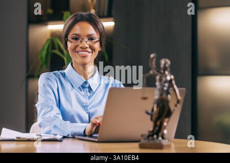 A businesswoman lawyer of African descent working at her desk with a statue of Lady Justice, making notes on paper. Stock Photo