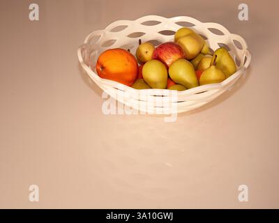 Still life of a white plastic lattice basket filled with fresh pears, apples, and an orange on a neutral surface in a home in Tuscany, Italy. Stock Photo