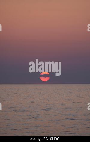 A vertical seascape with the golden sun hidden by clouds during a pink-purple twilight Stock Photo
