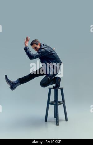 Energetic artist, young stylish man, leaping off stool in expressive motion, limbs extended against muted backdrop. Stock Photo