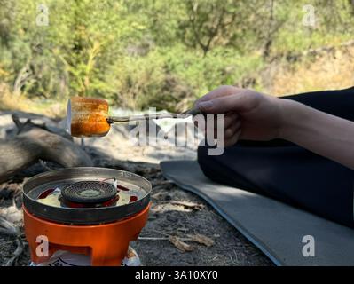 A close-up of a person roasting a marshmallow over a portable gas stove while resting in the mountains. The outdoor setting highlights a camping and t Stock Photo