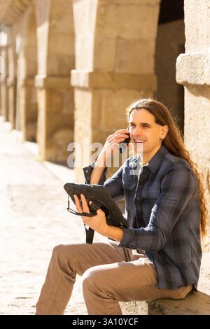 Smiling man talking on smartphone while using a rugged tablet outdoors. Lifestyle, technology. Stock Photo