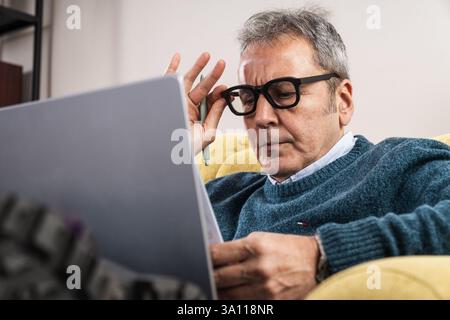 Close-up of a senior man wearing glasses, deeply focused while reading and analyzing documents on a laptop. Concept of business strategy, remote work, Stock Photo
