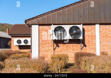 Two large electric fans are fixed on a red brick wall, supported by metal brackets. Their black blades contrast with the bright background. The Stock Photo