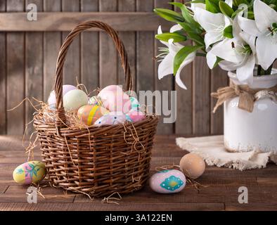 Decorated Easter eggs in a basket with white lilies on rustic wooden background Stock Photo