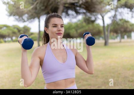 A Young Woman Energetically Lifting Weights in a Beautiful Park on a Sunny, Bright Day, Feeling Strong and Empowered Stock Photo