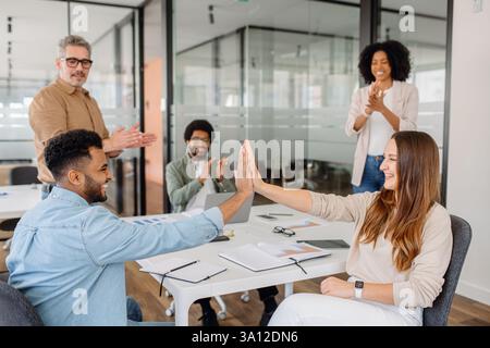 Two young professionals exchange a high-five at a desk, symbolizing teamwork and shared success. Colleagues celebrate an achievement in a modern office space, representing motivation and camaraderie. Stock Photo