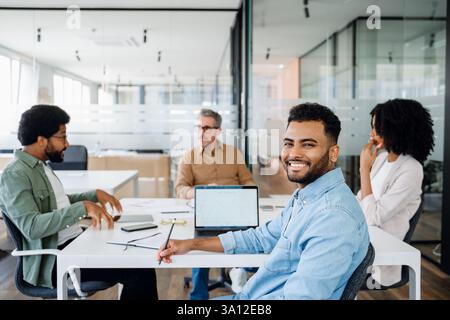 A diverse group of professionals engaged in a team meeting in a modern office. A senior executive leads the discussion while colleagues actively participate, taking notes and working on laptops Stock Photo