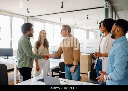 A professional business handshake between two colleagues in a modern office space. A diverse team watches in the background, emphasizing teamwork, collaboration, and partnership in a corporate setting Stock Photo