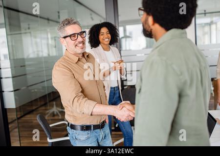 A professional handshake between two business colleagues in a modern office. Smiling coworkers observe, symbolizing agreement, teamwork, and successful partnership in a collaborative work environment. Stock Photo