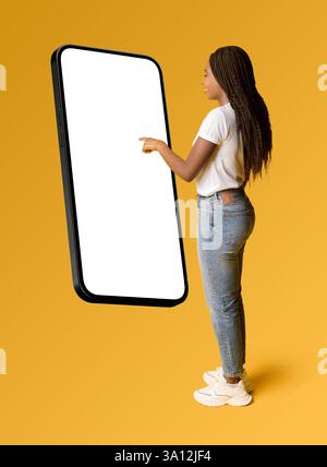 A young African American woman in casual jeans and a white t-shirt interacts with a giant smartphone mockup on a yellow background. Ideal for app promotions, mobile technology, and digital marketing. Stock Photo