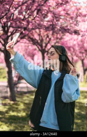happy woman listening music in headset outdoors taking selfie with sakura on background Stock Photo